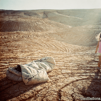 A young girl in pink top walks away across a desolate desert with cracked earth and a cru…