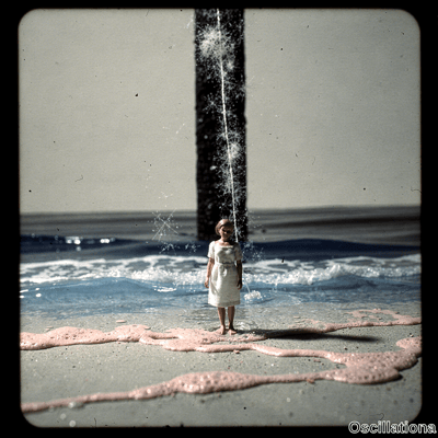 A young blonde girl in white textured dress stands barefoot at waters edge on beach looki…