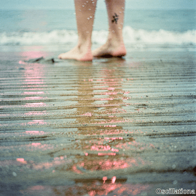 Bare legs and feet with black star tattoo on ankle standing in shallow beach surf as wave…
