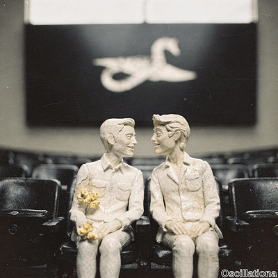 Sepia-toned diorama of two cracked porcelain figurines of young men on black leather thea…
