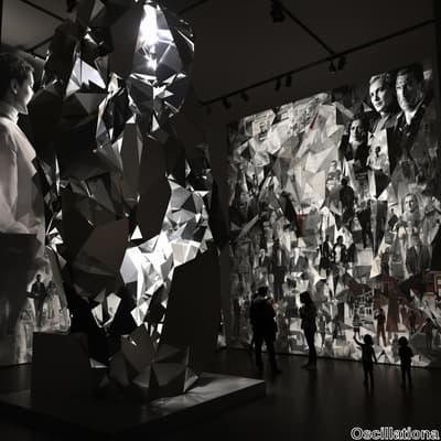 A large, abstract sculpture with reflective, angular facets stands in a dimly lit gallery…
